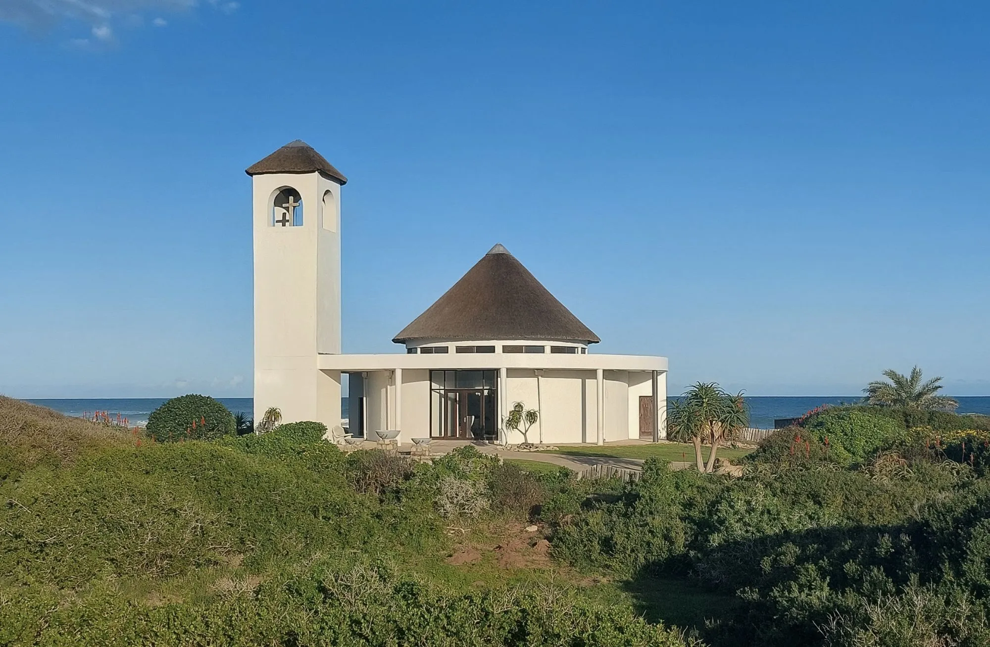 Monica's Chapel by Gourikwa: 'n wit kapel met 'n grasdak en 'n kloktoring, met die see daaragter.